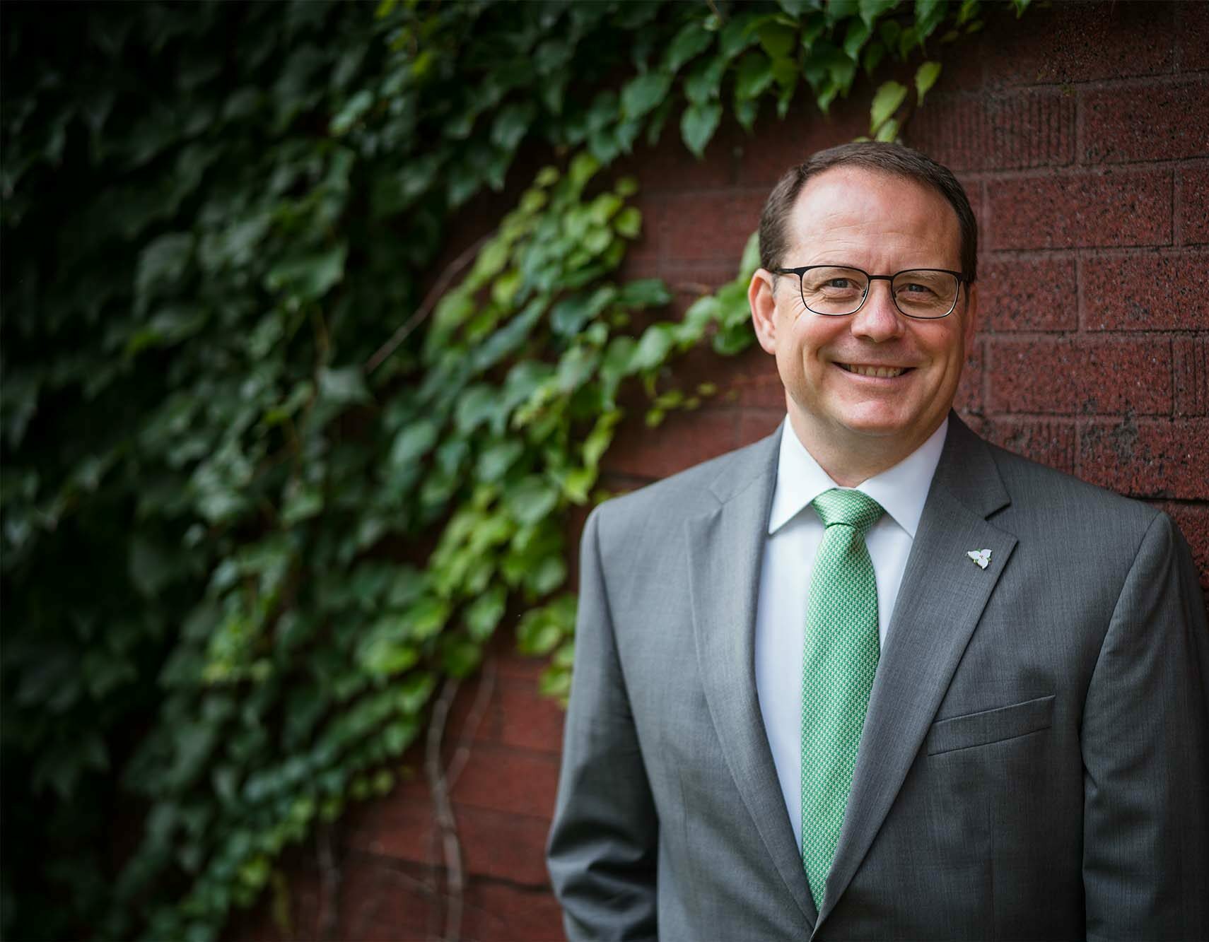 Mike Schreiner standing and smiling in a nice suit with. green tie.